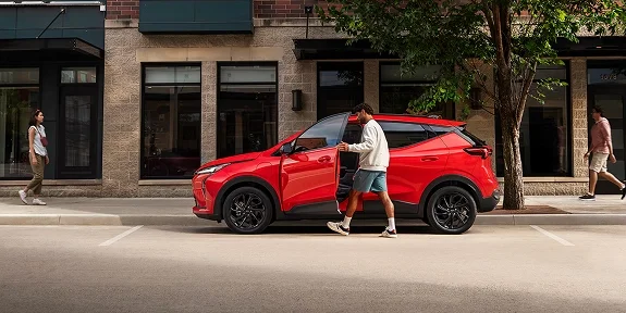 Side view of the 2027 Chevrolet Bolt RS in red on a city street with black wheels and roof rails visible